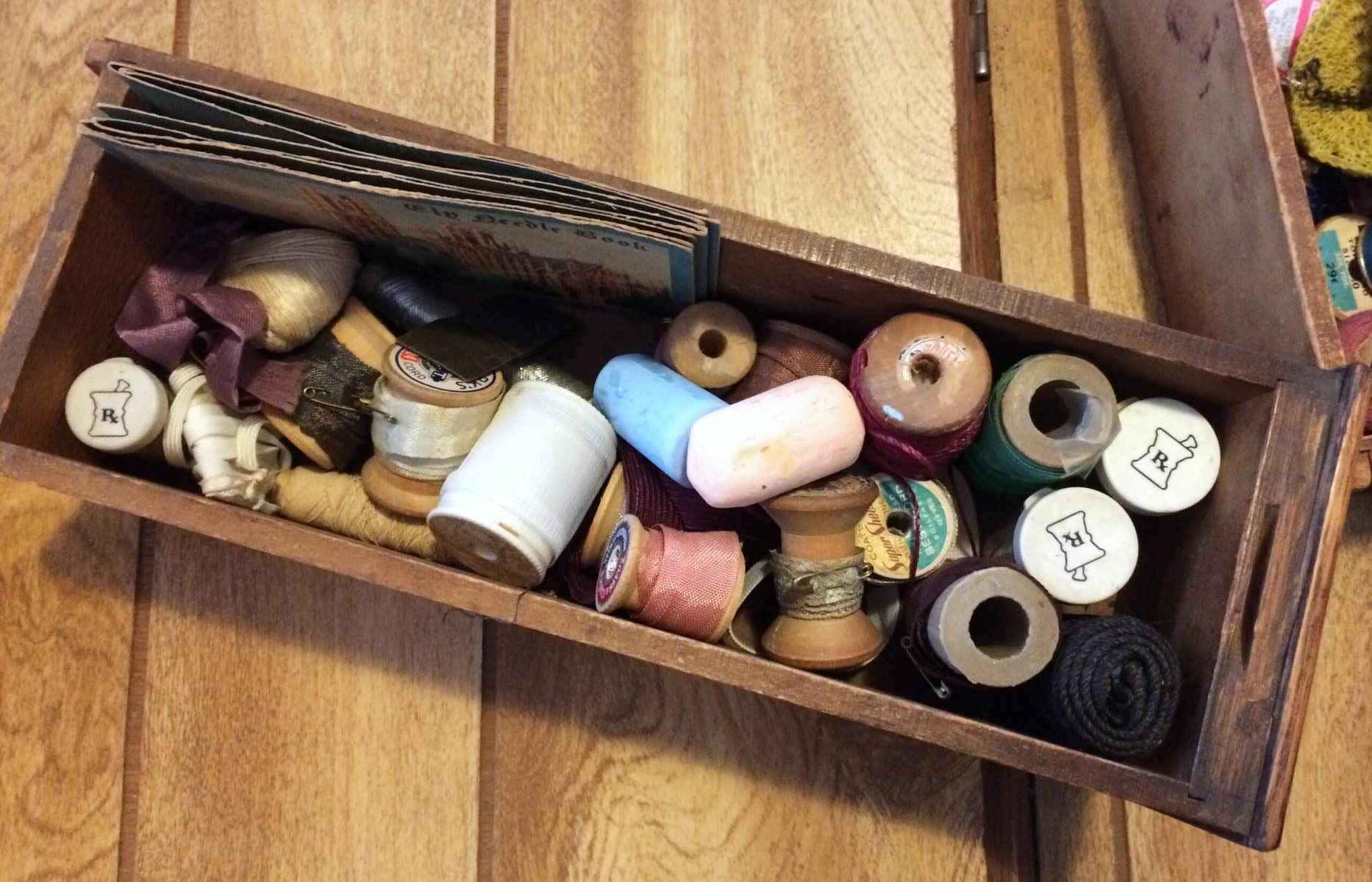 Photo description: A long, narrow drawer removed from its sewing table, seen from above and oriented at an angle horizontally, containing vingage spools of thread, pieces of tailor's chalk and more. 