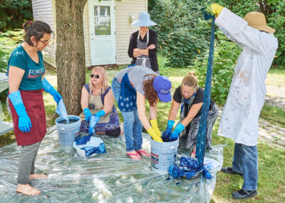 Six people outdoors where a plastic sheet is on the grass, with two buckets of blue liquid on it. Two women wearing gloves have their hands in one of the vats; another person is holding indigo-dyed fabric.