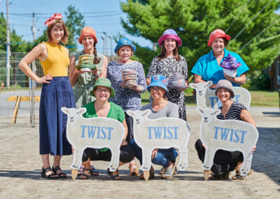Five women wearing hats, standing behind three women squatting bethind cardboard cutouts of sheep that say "TWIST" on them.