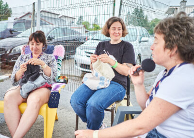Image description: Two women sitting and knitting, with a third, on the right ,speaking into a microphone.