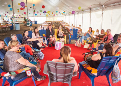Image description: People sitting in Adirondak chairs in a circle, in a large tent with paper lanterns overhead.