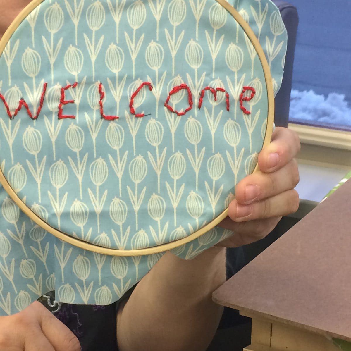 Image description: Embroidery hoop close-up, being held in one hand, with blue fabric with white flowers stretched in the hoop, with "WELCOME" embroidered in red thread.