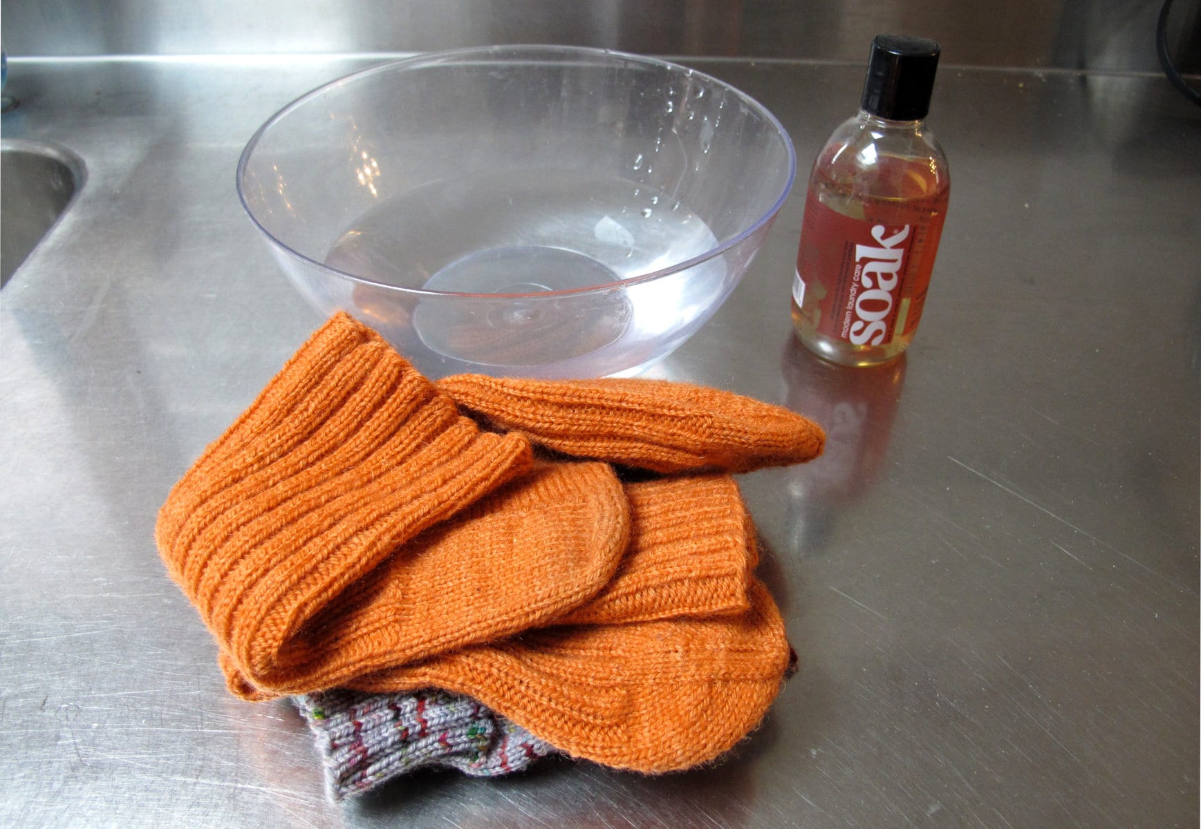 Image description: On a stainless steel countertop, an empty glass bowl, two pairs of folded socks, and a small bottle of Soak wash.