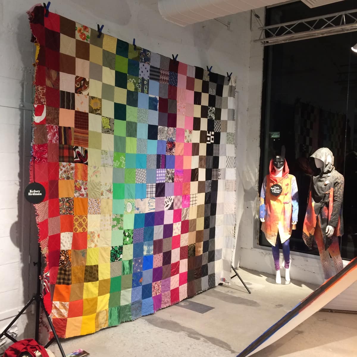 image description: a large, multicoloured quilt, hung on a wall in a gallery