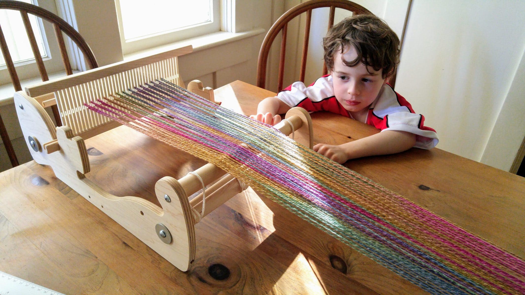 Image description: Small loom with yarn extending out from it, with a young child sitting at the table beside it.