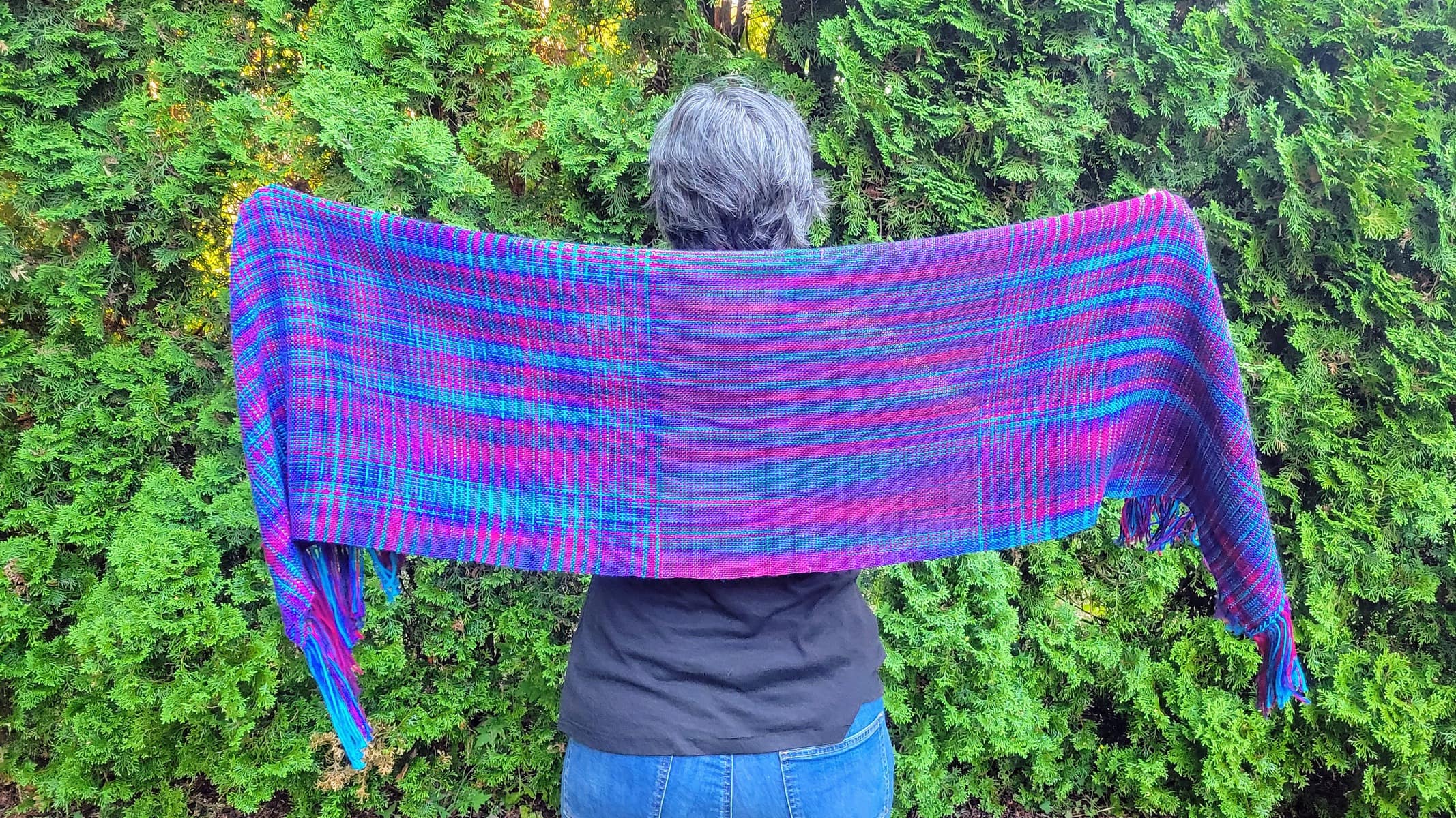 Image description: Grey-haired woman photographed from behind, holding a shawl out in both hands, in shades of blue and purple.