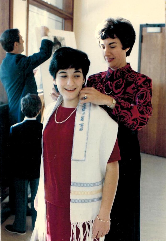 Image description: An adult woman adjusts the neck of a tallit on a young teenage girl.