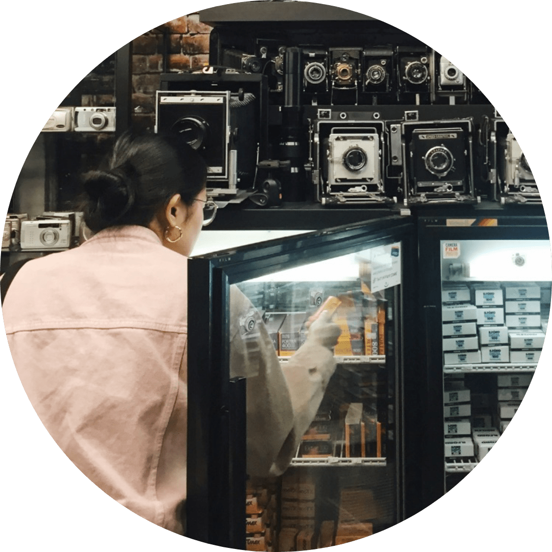 Image description: Woman in pink jacket is reaching for an item in an open glass case. Above the case are many vintage film cameras.