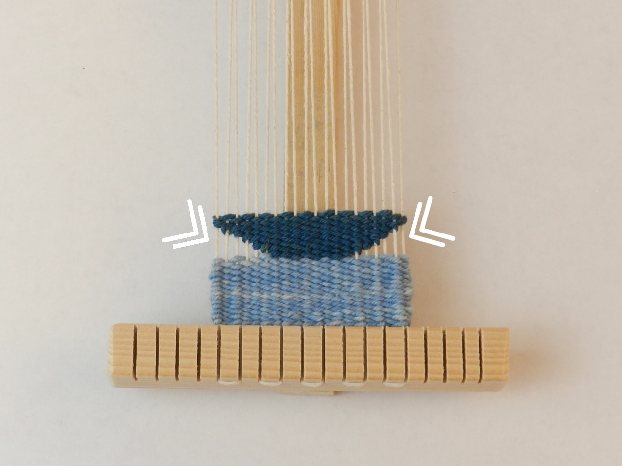 image description: a narrow warp of white yarn has about an inch of light blue weaving at the bottom, with navy weaving in the shape of an inverted trapezoid on top of the light blue, with a small triangle unwoven at either side. Arrows point to the unwoven triangles.