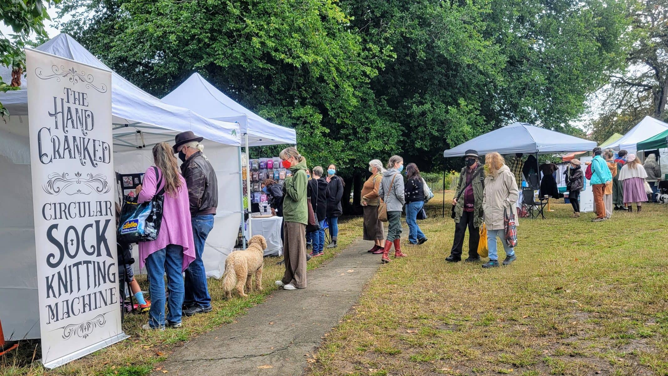 Image description: People milling around at an outdoor festival. A vertical sign on the left reads, "The Hand Cranked Circular Sock Knitting Machine."