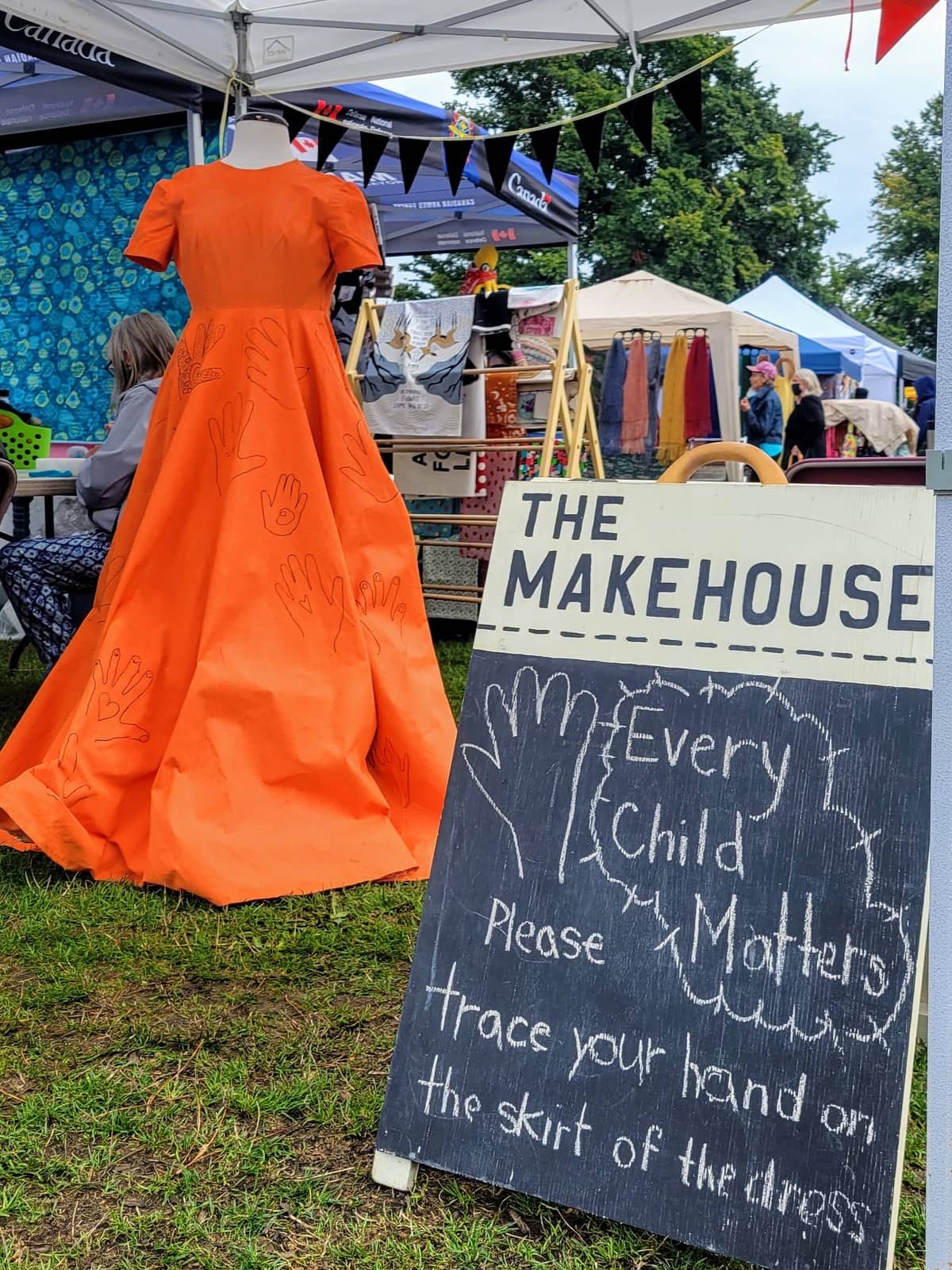 Image description: An orange gown with hands drawn on the skirt is on a dressform next to a chalk sign reading "Every child matters. Please trace your hand on the skirt of the dress."