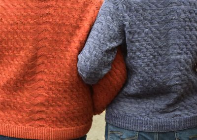 Image description: Man in orange sweater and woman in blue sweater, standing side by side with linked arms, seen from behind.