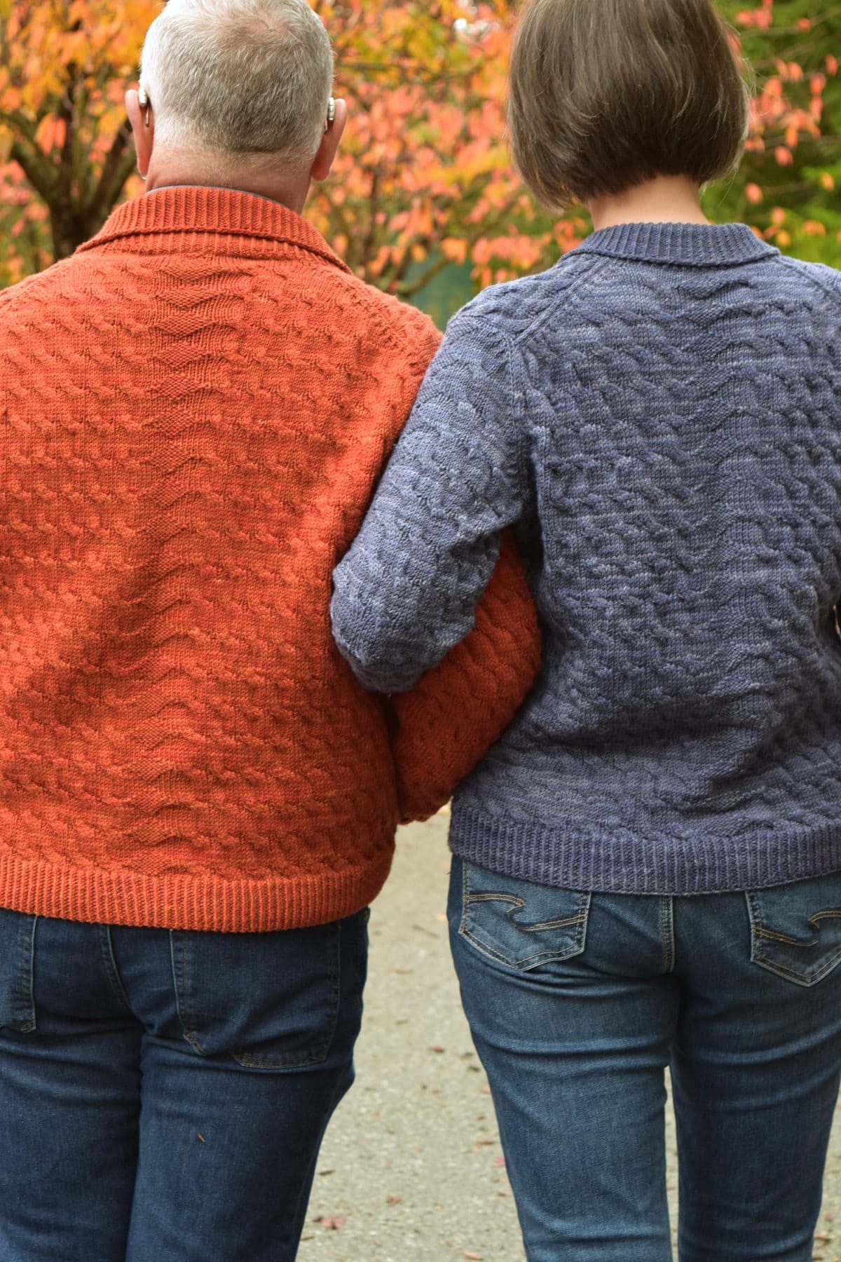 Image description: Man in orange sweater and woman in blue sweater, standing side by side with linked arms, seen from behind.