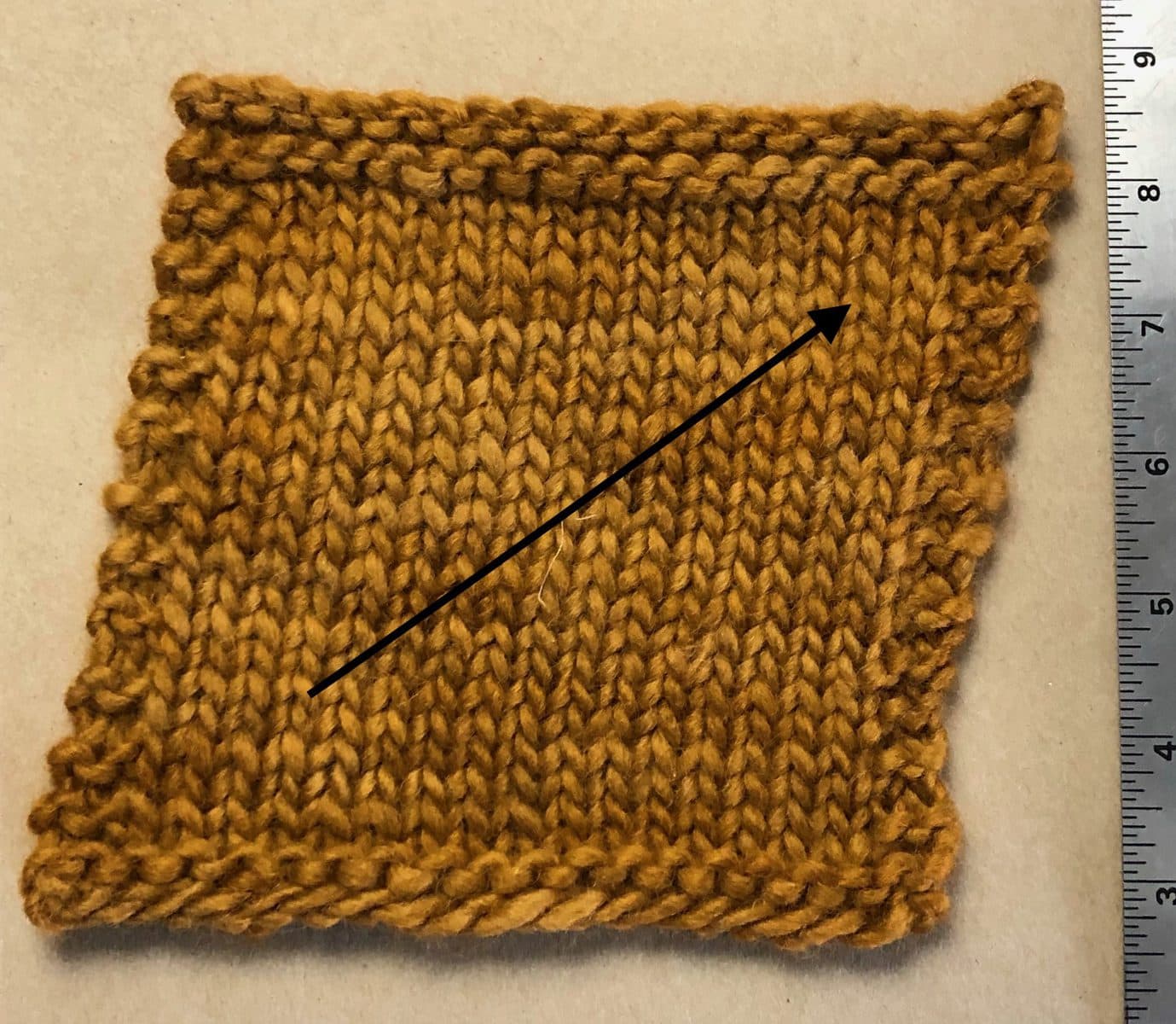 image description: a swatch of stockinette stitch, with a noticeable right-leaning bias; an arrow is superimposed on the image to draw attention to the lean