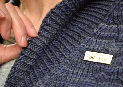 Image description: Gold badge with black writing that says "She/Her" is pinned to a blue-grey cardigan.