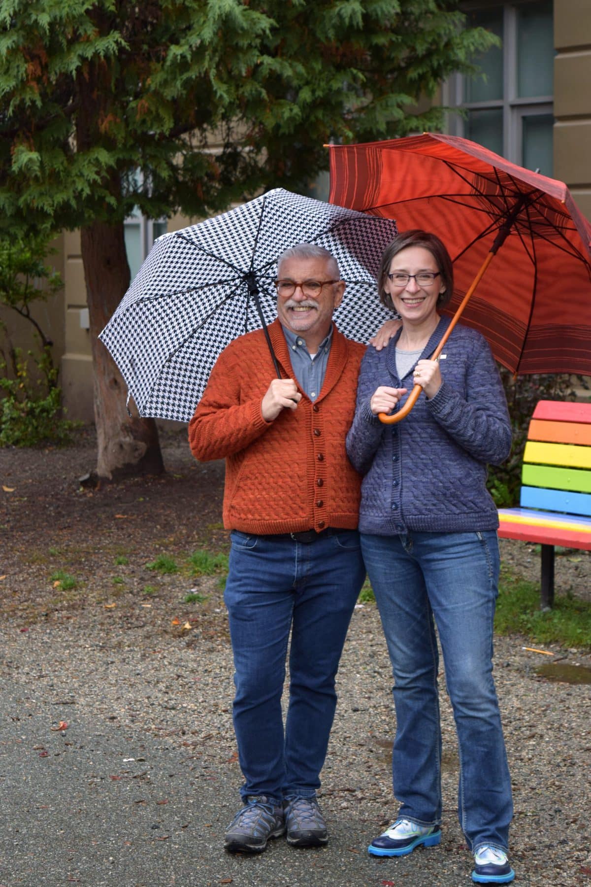 Image description: Man in orange cardigan and woman in blue cardigan, each holding an umbrella, stand in front of a rainbow bench.