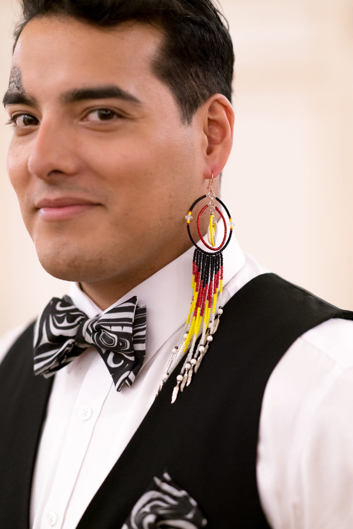 image description: the head and shoulders of a man in three-quarters profile; he is wearing a single beaded earring in his left ear, a bow tie in a black and white pattern associated with indigenous art, and he has a small tattoo of a similar pattern over his right eyebrow