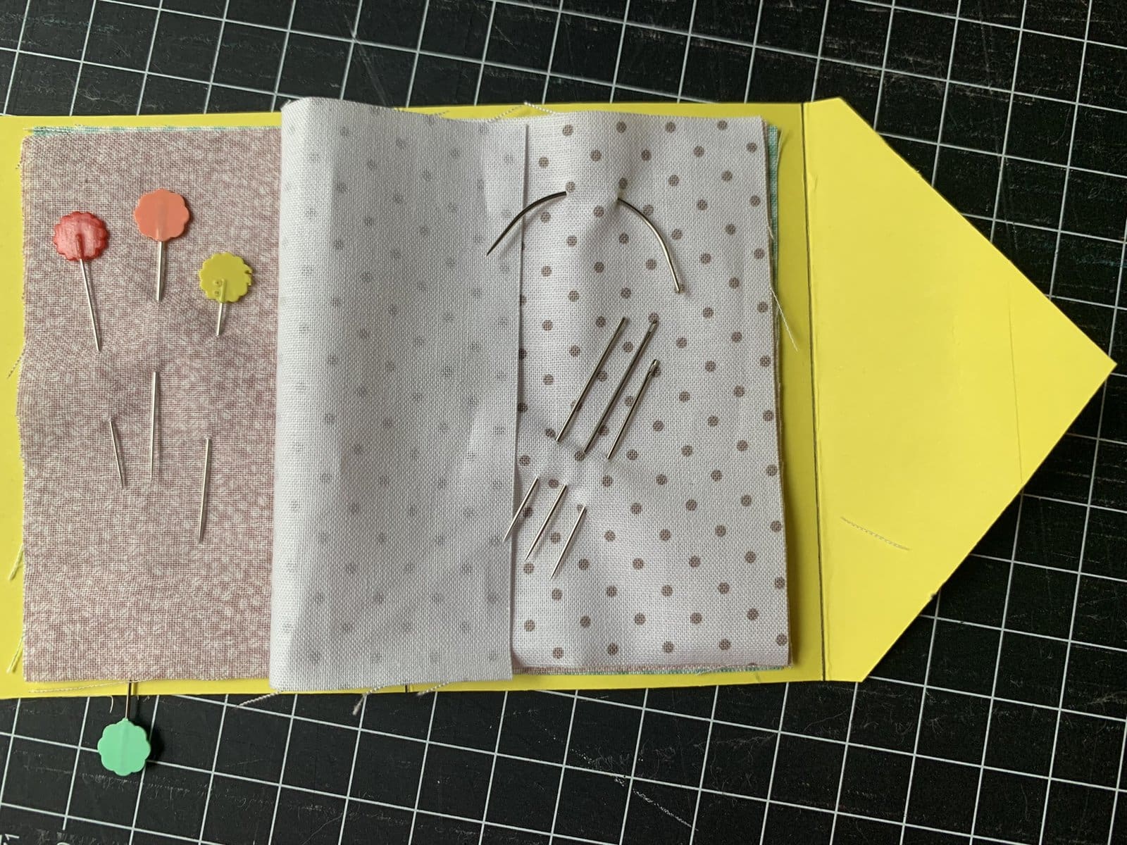 image description: a needle book, open on a table; it's made of a cardstock cover and fabric pages, and a few needles and pins are clipped into the pages