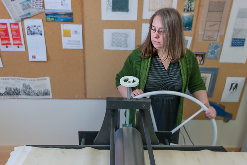 Image description: White woman with shoulder-length hair wearing a green cardigan over a black dress holds a large white metal wheel on the side of a letterpress machine.