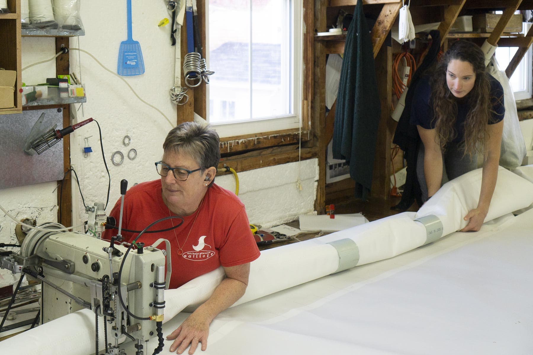 image description: two people working on an enormous piece of white fabric - one is operating a sewing machine, the other is holding a roll of the fabric, guiding it and supporting its weight