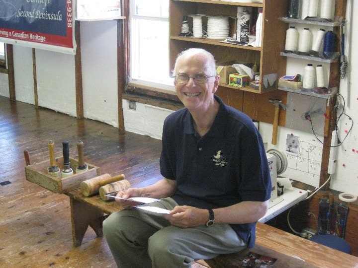image description: an older white man sits on a bench in a workshop, he is looking directly at the camera, a broad smile on his face