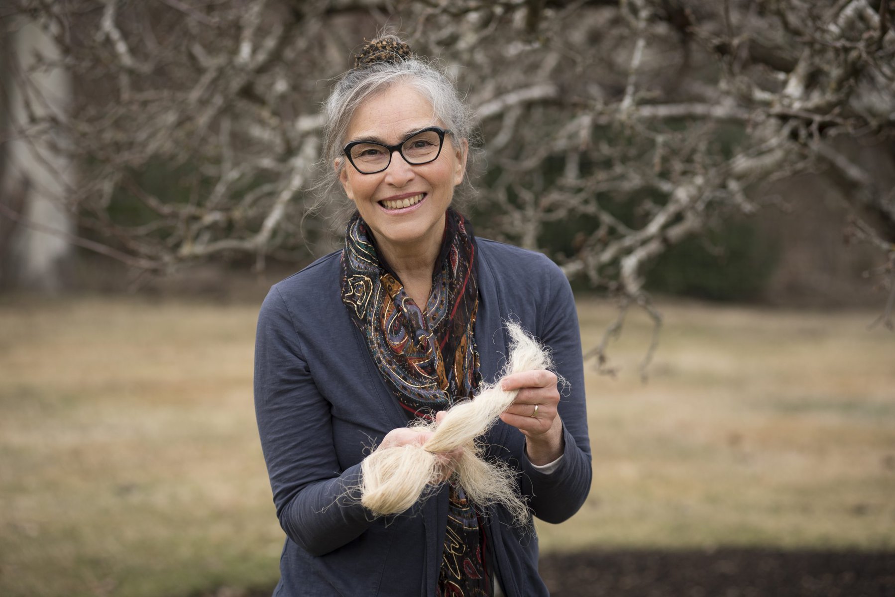 image description: a portrait of a white woman; she wears glasses and her greying hair is pulled up into a bun; she is holding strands of straw-coloured fibre